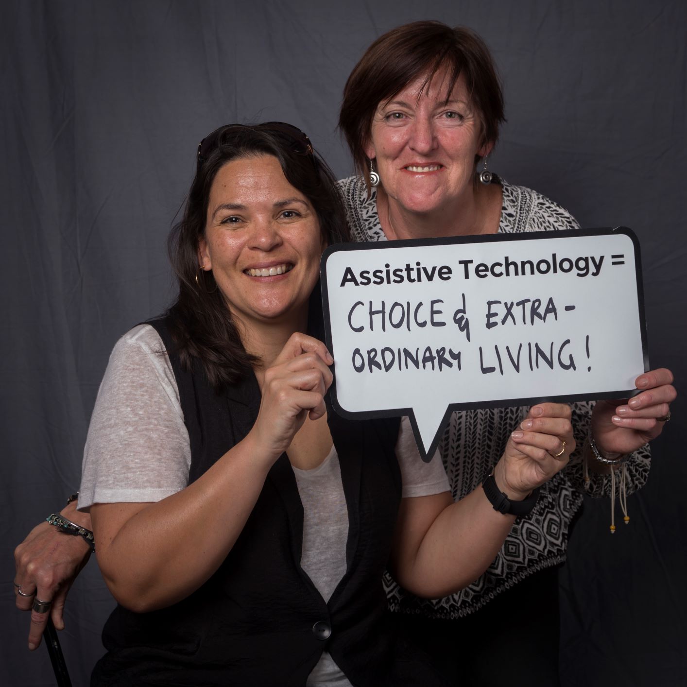 two woman holding up a speech bubble sign that reads assistive technology = choice and extra ordinary living