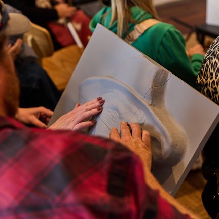 Person wearing a red and black chequered shirt touching a grayscale 2.5D photograph of a mans face, hat and clothing, seated indoors with others nearby