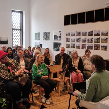 People seated in a gallery style room listening to a male speaker in a green shirt, with photographs displayed on the walls