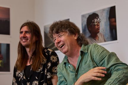 Two people seated indoors during a discussion, with several photographs displayed on the wall behind them.
