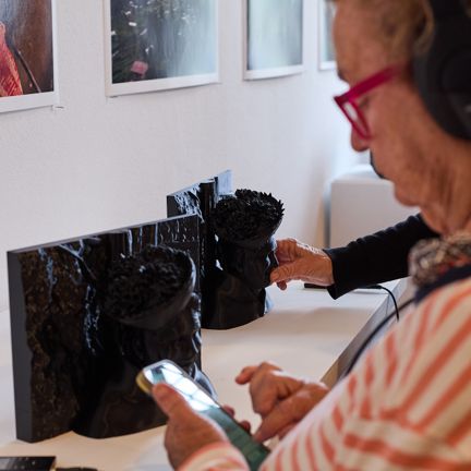 Person using headphones and a handheld device while exploring black tactile 2.5D photograph on a table.