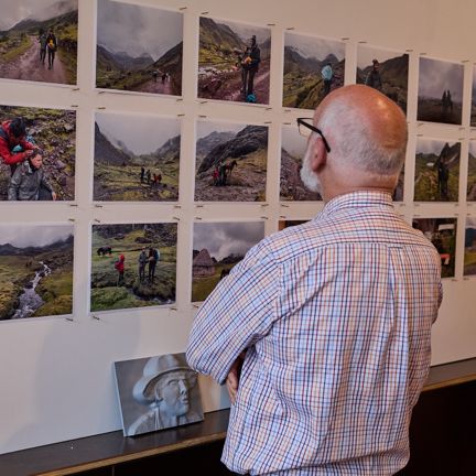 Person viewing a display of multiple landscape photographs arranged in a grid on a gallery wall, with a grayscale 2.5D photograph below.