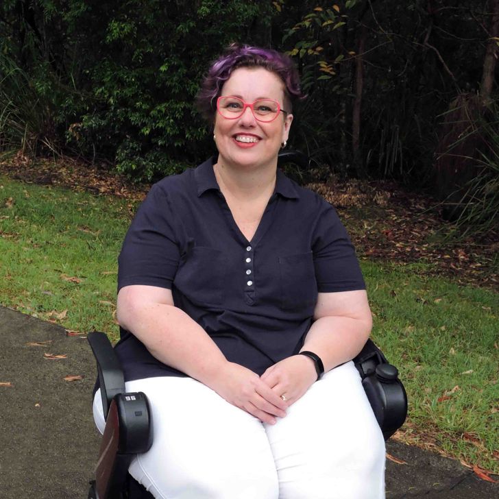 A smiling woman wearing red glasses and a navy and white outfit, using a powered wheelchair