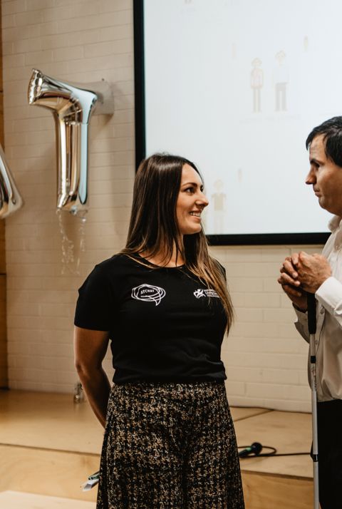 A woman and a man are standing talking, with a pair of balloons spelling out AT in the background