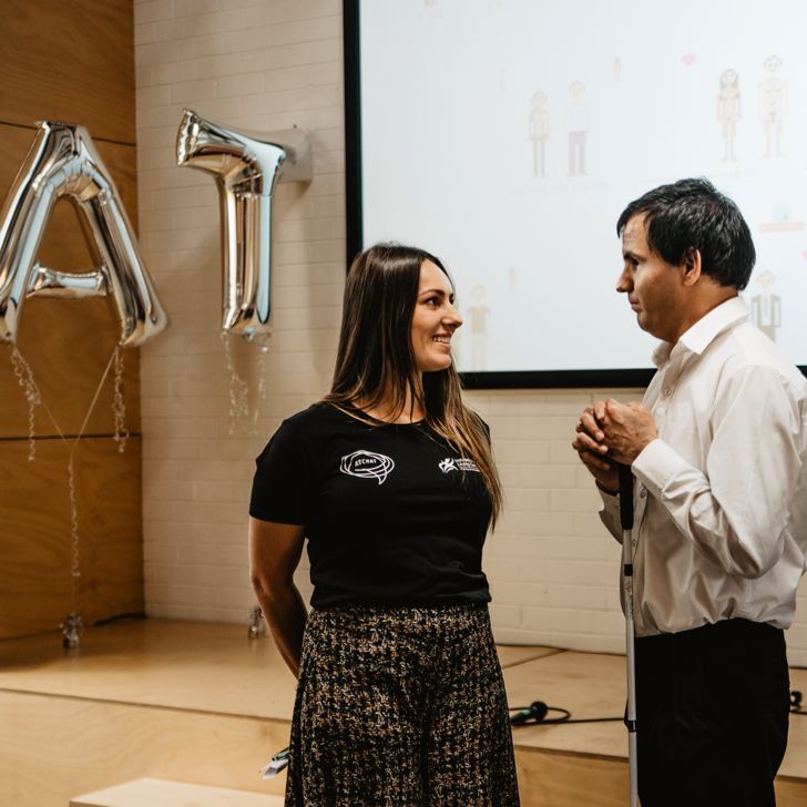 A woman and a man are standing talking, with a pair of balloons spelling out AT in the background