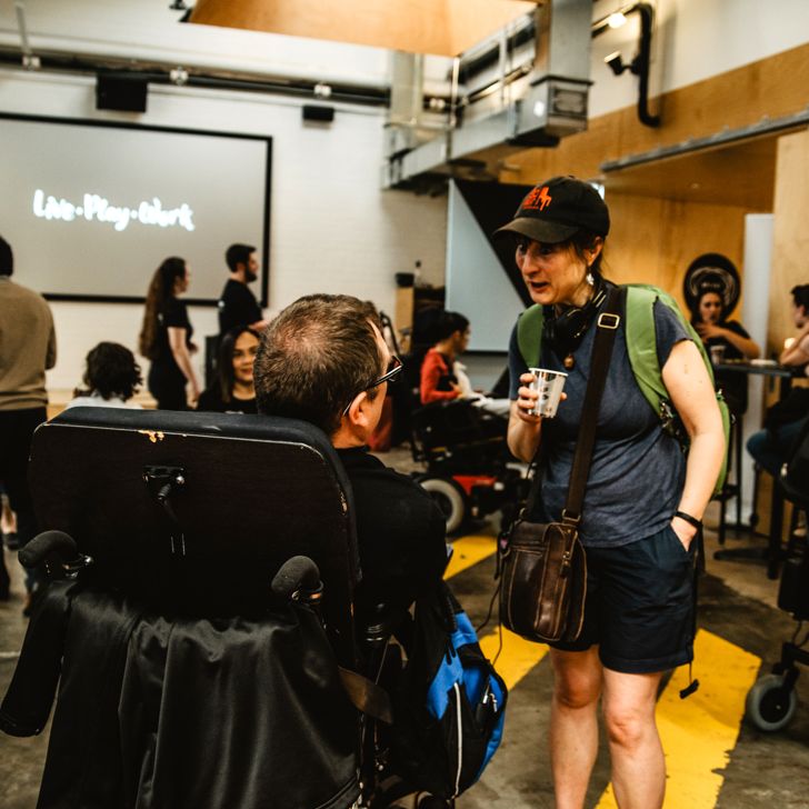 A man in a wheelchair talking to a woman who is standing, in a room full of people and the words Live, Play, Work on a screen in the background