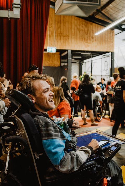 Man in electric wheelchair smiling at another man standing in front of a crowd.