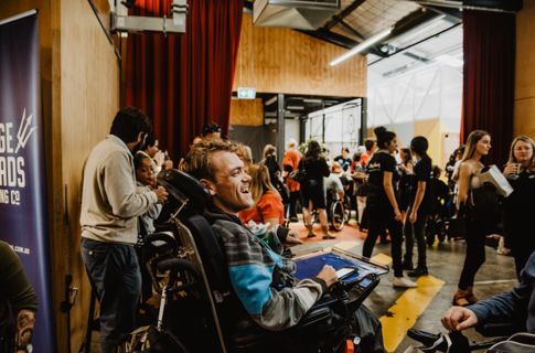 A man in an electric wheelchair faces away from the camera and smiles in conversation with another man in an electric wheelchair. They face a crowd of people. 