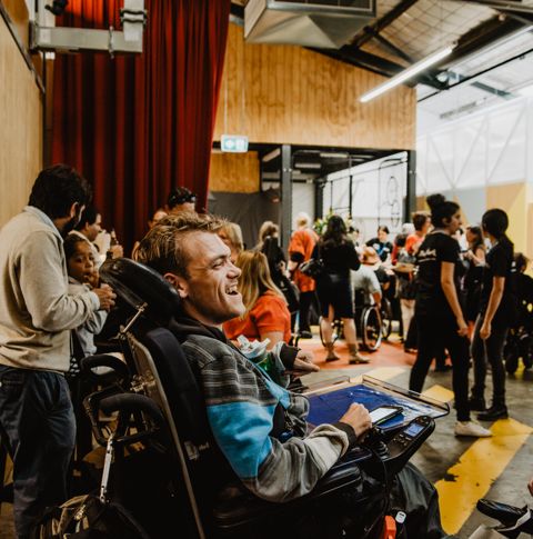 A man in an electric wheelchair faces away from the camera and smiles in conversation with another man in an electric wheelchair. They face a crowd of people. 