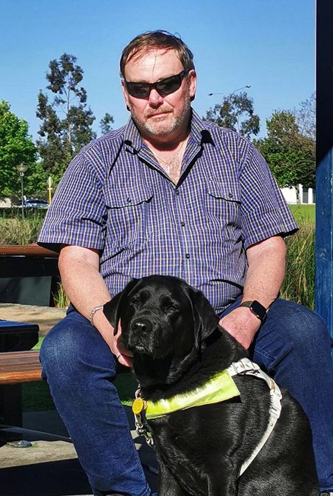 A man in sunglasses sits with his assistance dog
