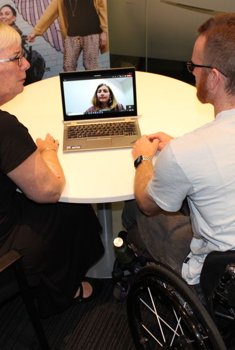 A woman and a man sit at a table looking at a laptop, the man is sitting in a wheelchair