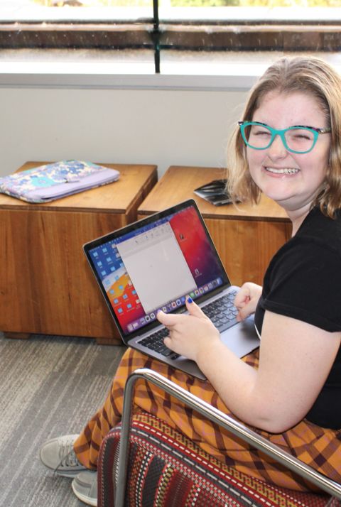 A young person sits inside using a laptop and smiling widely