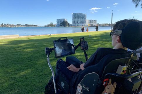 A young man in a wheelchair at a river side, facing the water on a sunny day