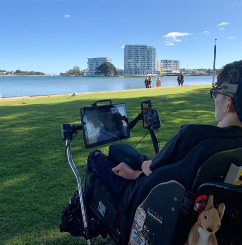 A young man in a wheelchair at a river side, facing the water on a sunny day