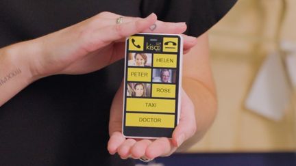 Hands holding a KISA Phone with large high-contrast simplified buttons and pictures.