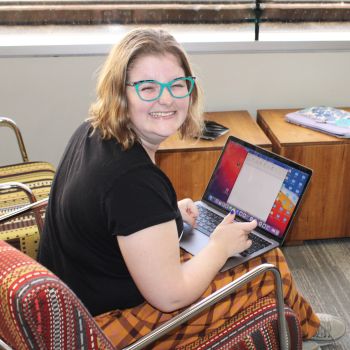 A young person sits inside using a laptop and smiling widely