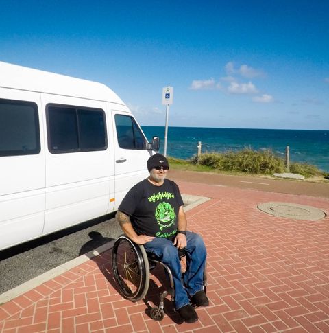 Mark sitting next to the white van on a sunny day at the beach.