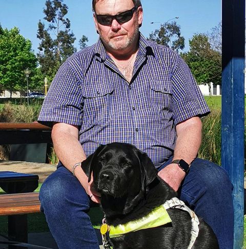 A man in sunglasses sits with his assistance dog