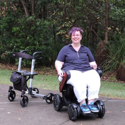 A woman sits, smiling in a power wheelchair outside, with a rollator to her right