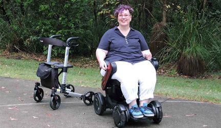 A smiling woman sits in a powered wheelchair, with a rollator on the side