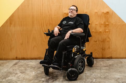 Nick in wheelchair sitting against wooden wall.