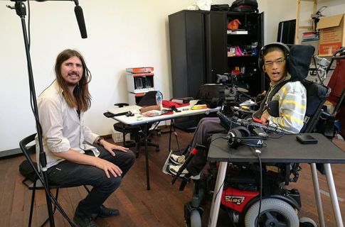 Two men in an audio studio, one of whom is in a power wheelchair