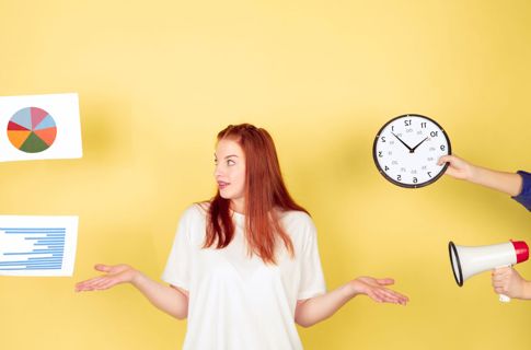 Photo of a woman looking confused with people's hands extending towards her holding a megaphone, clock and documents