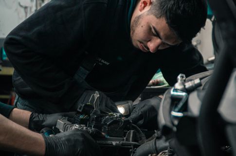 A young man leans over to look closely at the engine of a car, while someone else's hands make adjustments