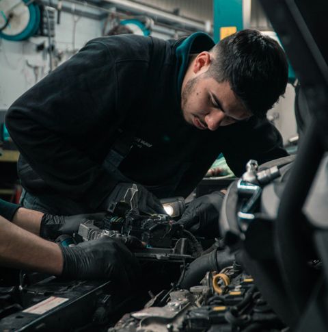 A young man leans over to look closely at the engine of a car, while someone else's hands make adjustments