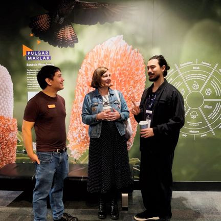A woman and two men stand in front of a backdrop with Australian native flora and fauna, talking and listening with interest
