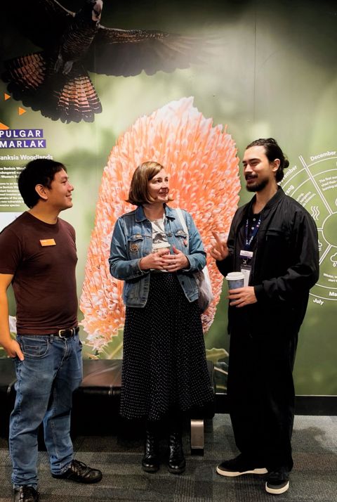 A woman and two men stand in front of a backdrop with Australian native flora and fauna, talking and listening with interest