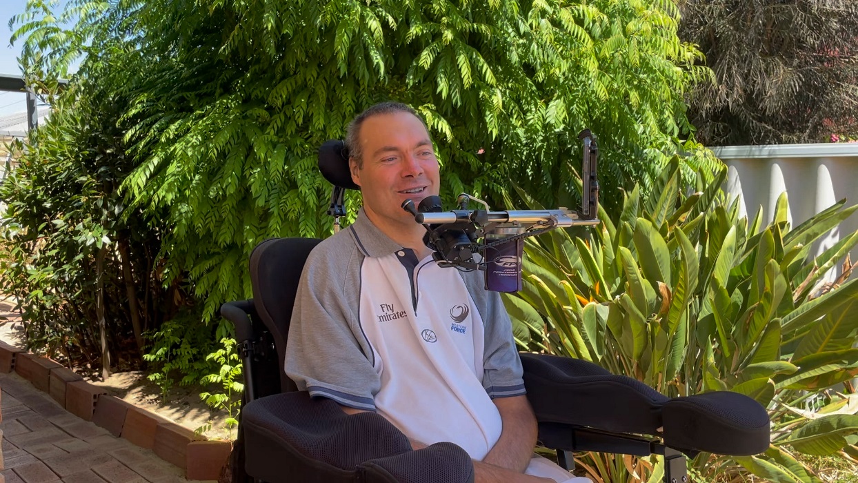 A man is seated outside in his powered wheelchair smiling at the camera. 