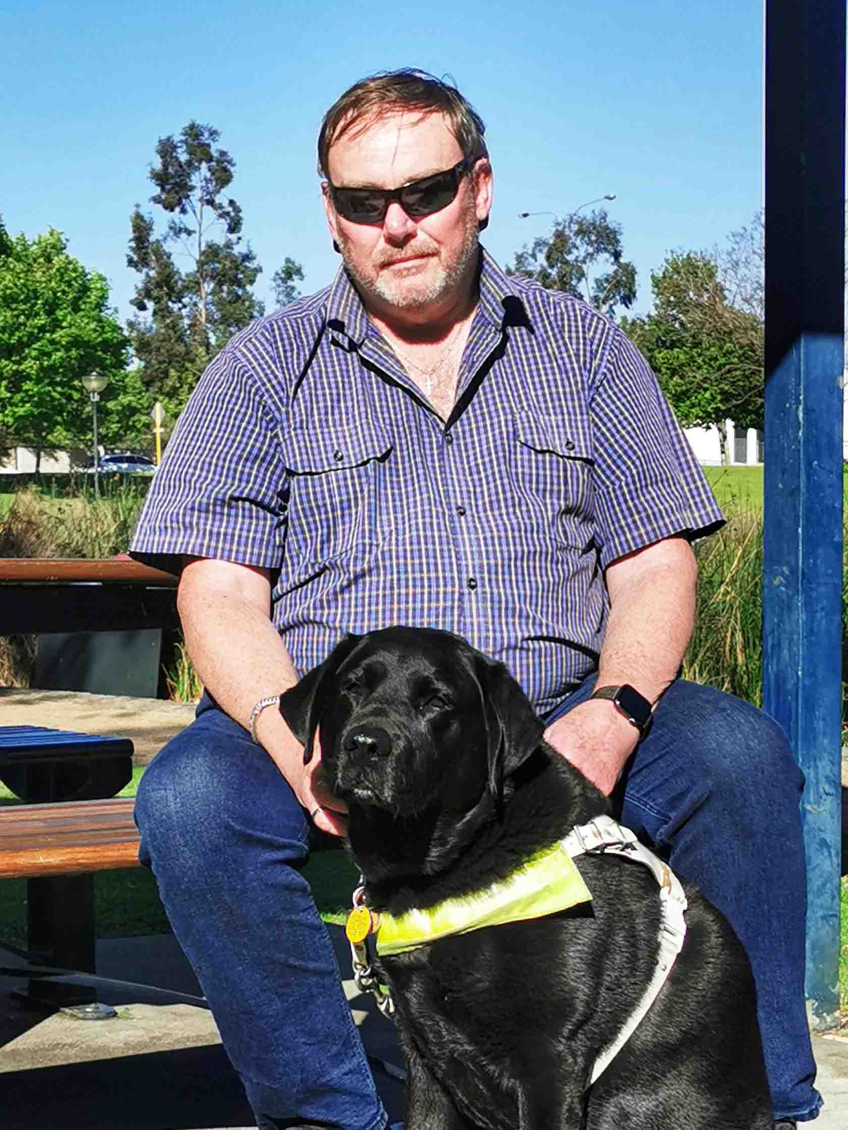 A man wearing sunglasses is seated outside with his guide dog