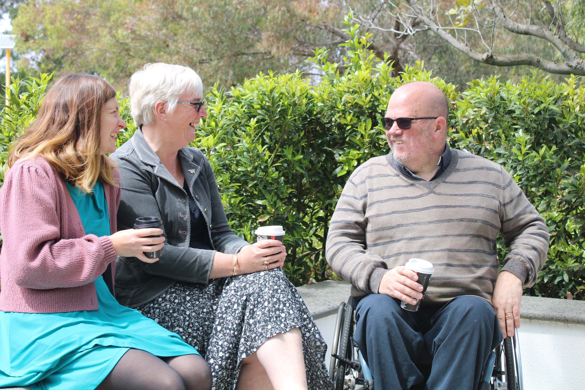 Two women are seated on a low stone wall beside a man seated in his wheelchair. They are all holding cups of coffee while smiling and laughing.