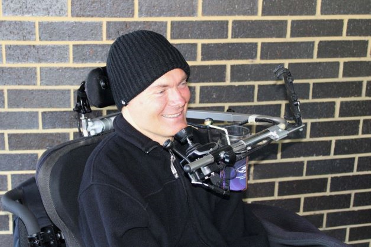 A man sits in his powered chair against a brick wall and smiles off to the right. A man sits in his powered chair against a brick wall and smiles off to the right.