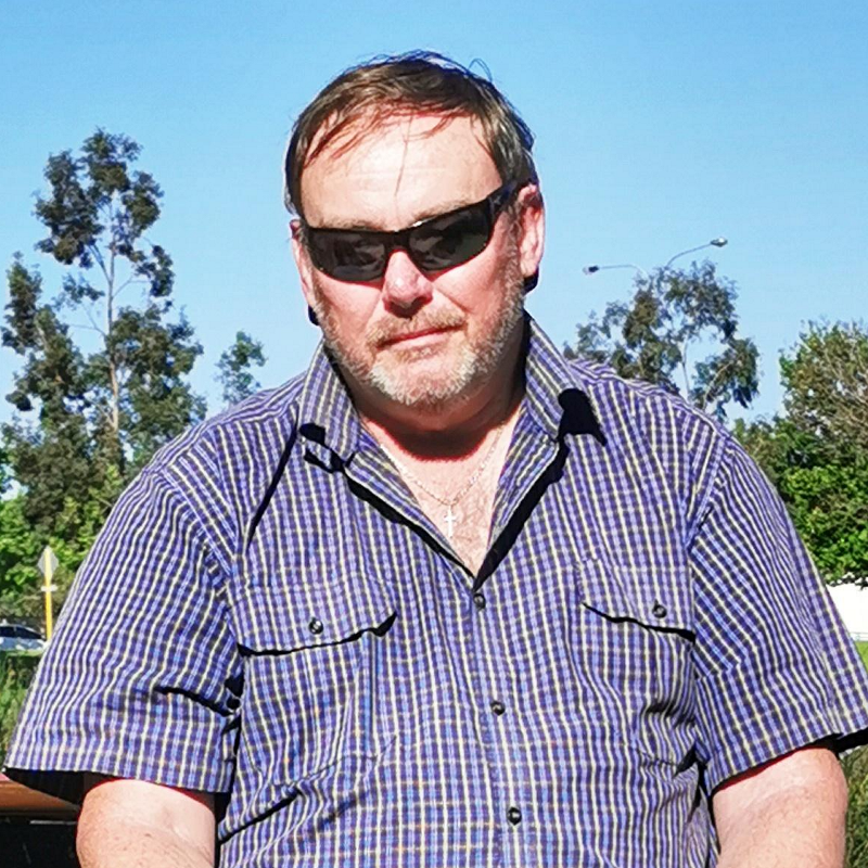 A man is seated at a picnic table wearing sunglasses, his guide dog is seated at his feet.