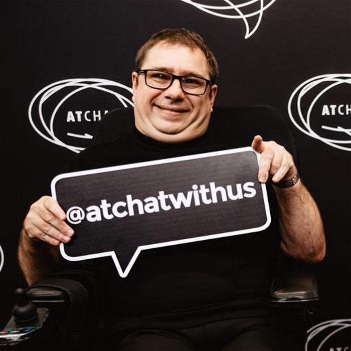 nick man sitting in wheelchair holds a sign that reads @atchatwithus