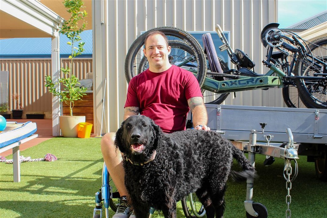 smiling man wearing red tshirt sits in a wheelchair patting a black dog in backyard