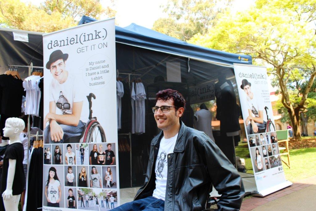 Daniel is at the markets seated in front of his stall with which has his range of t-shirts.  There are two promotional banners to each side of the stall showing a photo of himself and some of his t-shirts.