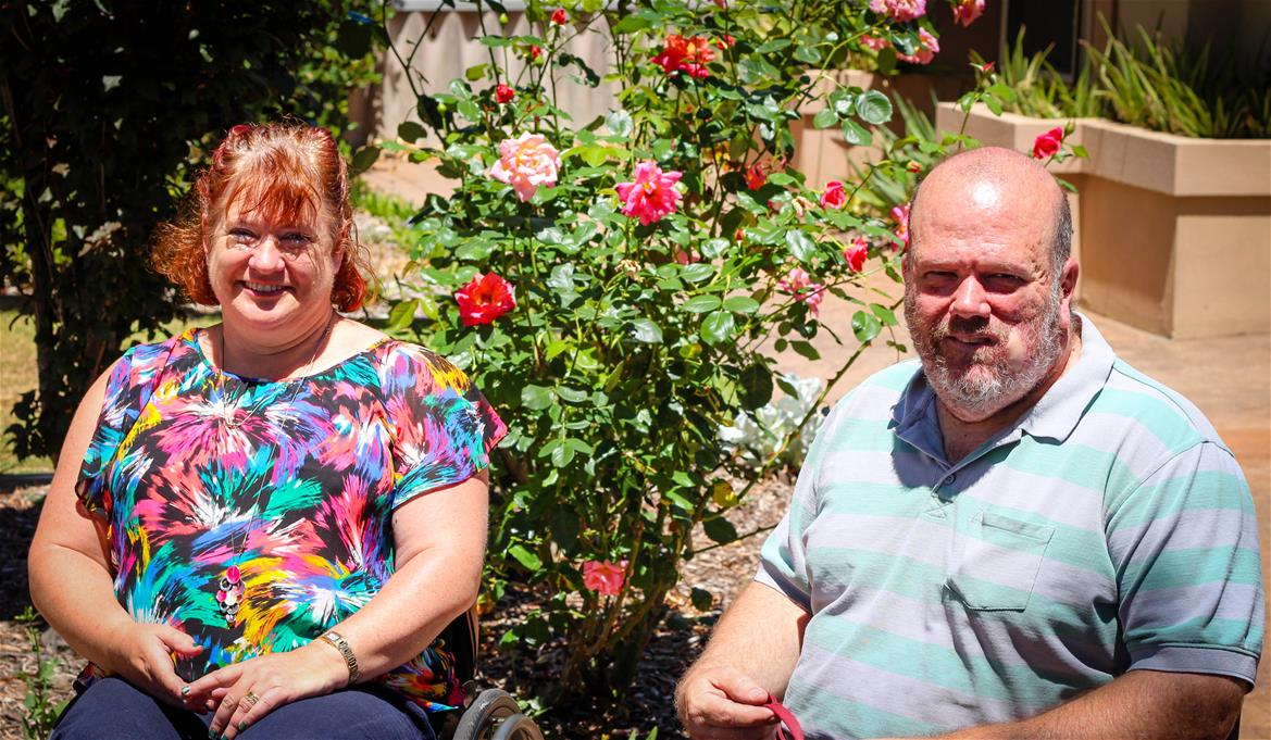 woman in floral top and man in striped polo sit in wheelchairs outside in garden