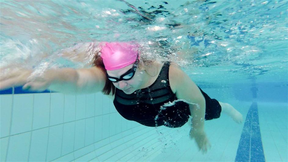 woman wearing black bathers, goggles and a pink swimming cap swims freestyle