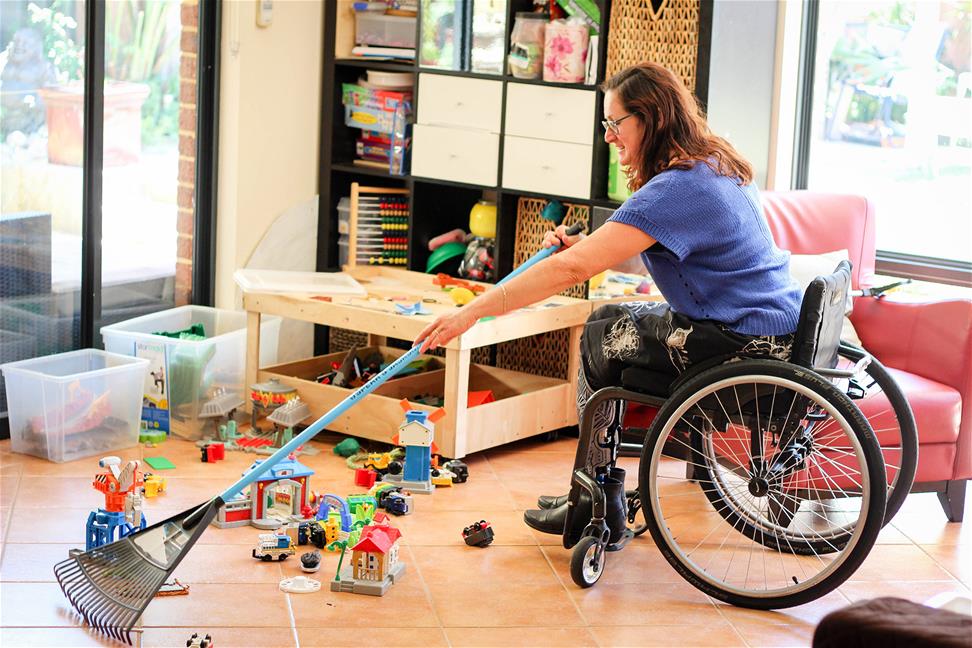 woman in wheelchair uses a rake to clean up toys on floor