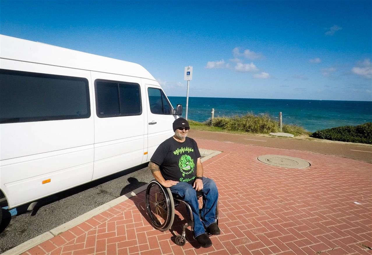 man wearing black tshirt and beanie sits in wheelchair in front of white van parked at the beach