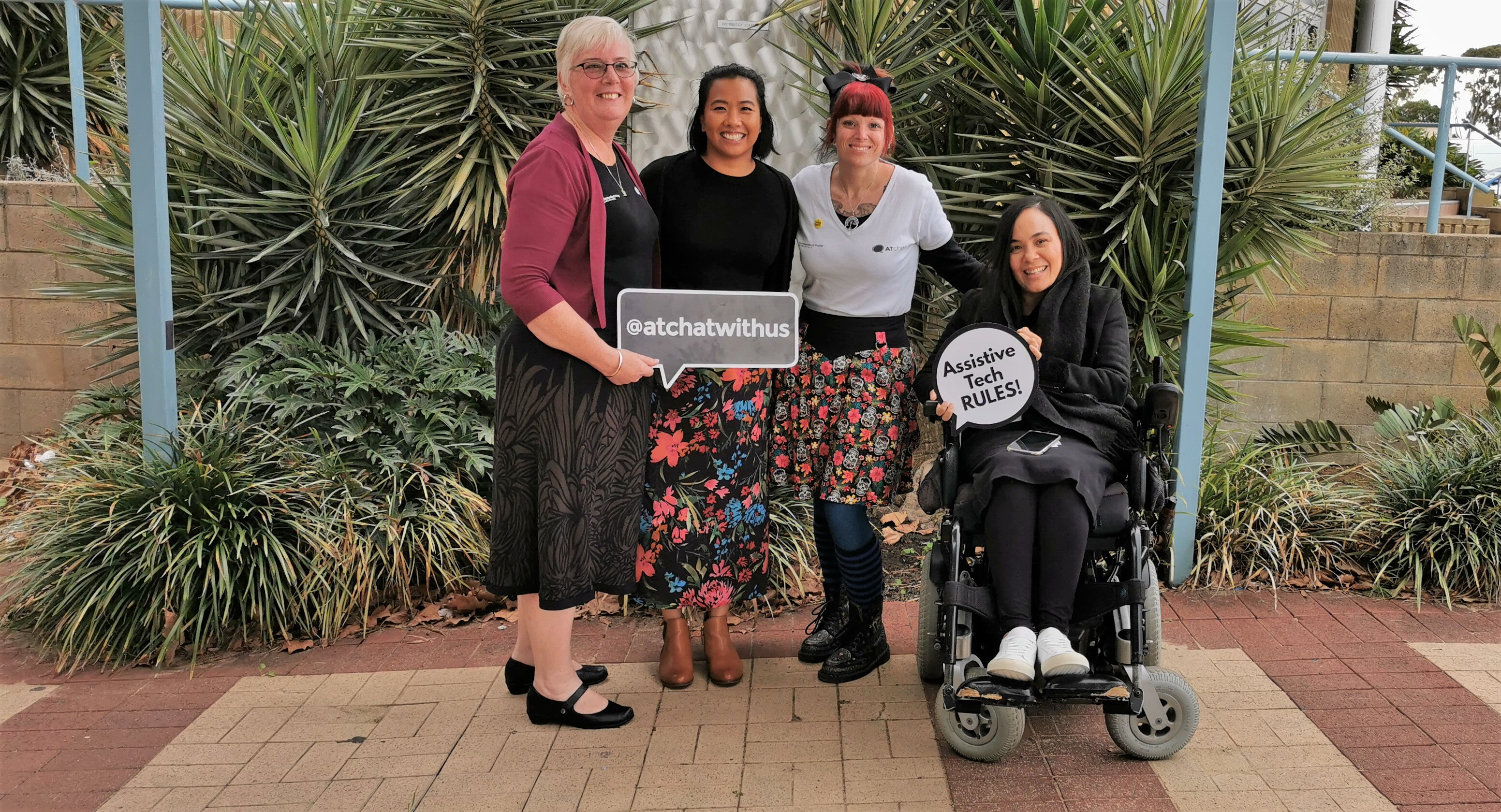 Four women gather together in front of the garden and smile holding AT signs.