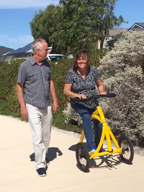 Man walks beside a womand who is riding the Alinker walking bike on a track in a park.  They are smiling and chatting with each other. 