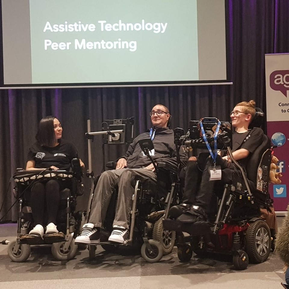 One woman and two men are seated in their wheelchairs on stage in front of a screen showing the AT Chat banner with their names underneath.