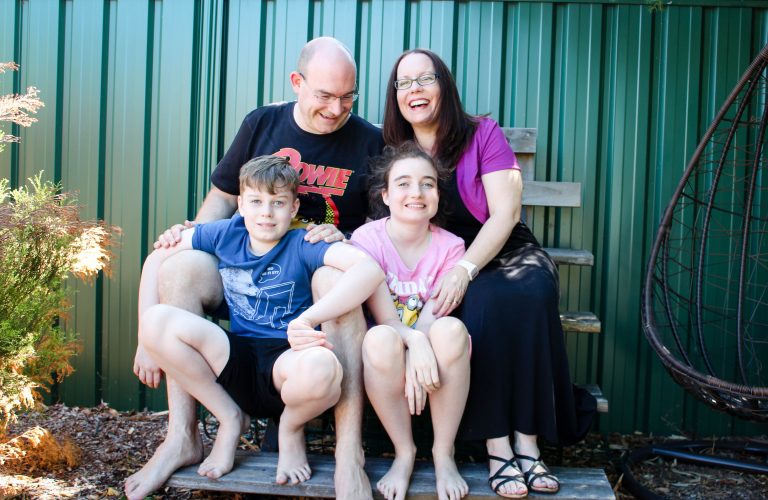 The Bolger family are seated together on a bench in their yard and smiling and laughing. Text overlay says blog post life as a family of four on the Autism spectrum