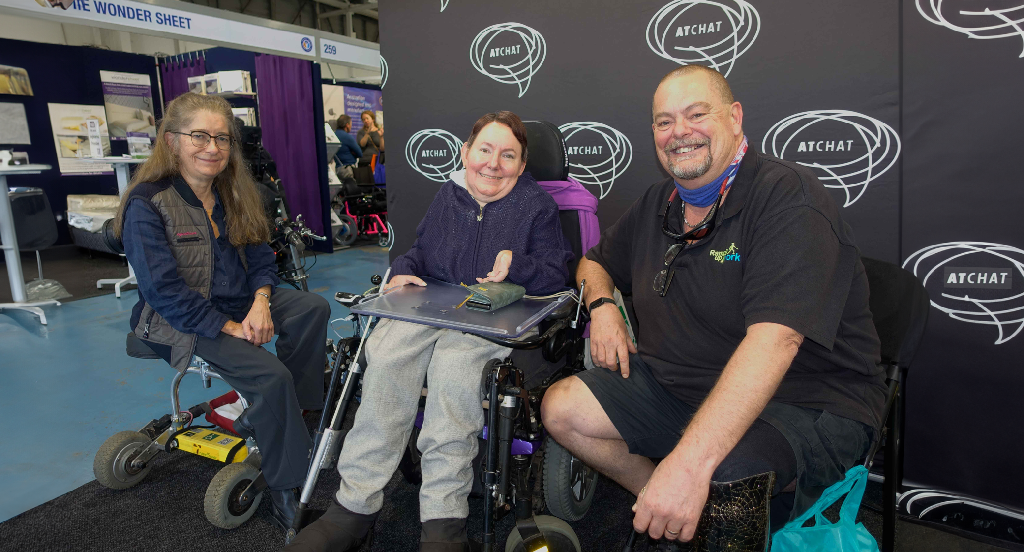 Fluer-Melanie-and-Andrew Perth ATSA Two women and a man are seated beside each other in front of the AT Chat banner smiling at the camera. One is seated on her travel scooter, one in her powered wheelchair and the man on a seat with his prosthetic leg stretched out in front of him.