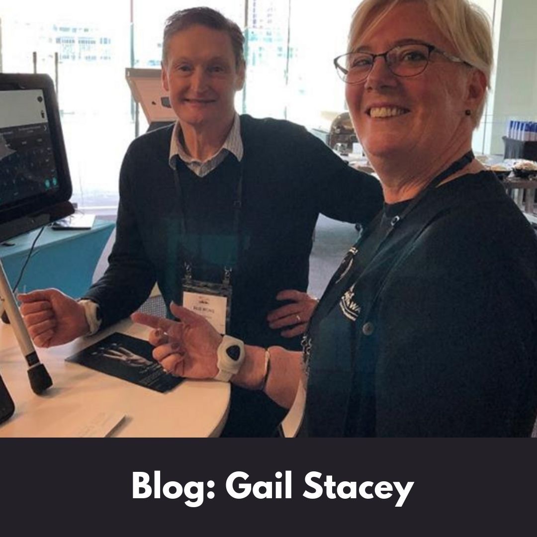 Woman wearing glasses is standing in front of a screen with her head turn towards to the camera. she has a neuro node sensor on her wrist. There is a man standing beside her also smiling at the camera. White text on black background says; Blog: Gail Stacey