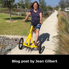 A woman wearing a blue top and denim shorts smiles at the camera as she sits on a yellow three wheeled walking bike on a path near a park on a sunny day. White text on a black background says AT Blog by Jean Gilbert: Funding my Alinker Walking Bike.